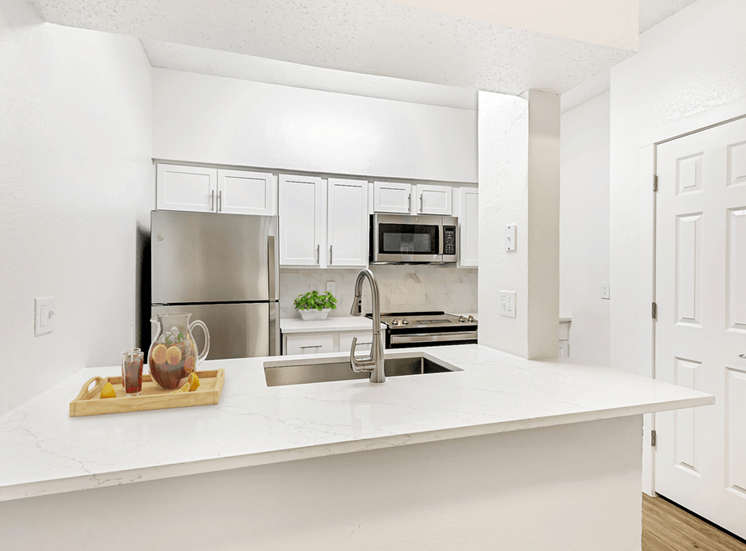 Virtually staged kitchen with white quartz countertops, white cabinetry, stainless appliances, goose neck faucet with sprayer and serving tray with pitcher of tea